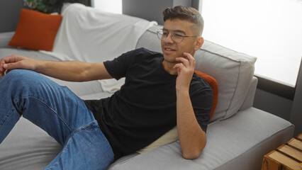 Fototapeta premium Young hispanic man relaxing on a couch in a stylish living room with modern decor at home, showcasing a comfortable and inviting interior setting.