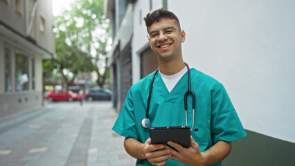 Young hispanic male nurse smiling outdoors holding a tablet on an urban street wearing scrubs and stethoscope suggesting healthcare professionalism.