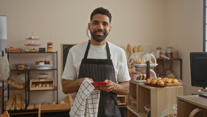 Young man in bakery shop smiling while holding dish towel in modern, indoor setting with breads and pastries displayed in the background