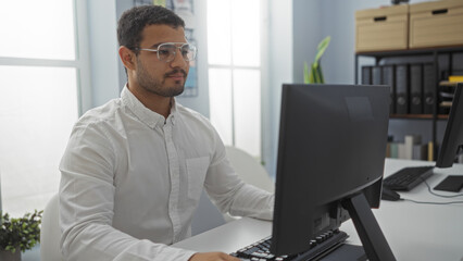 Hispanic man working on a computer in an office, focused and wearing glasses, with a modern indoor...