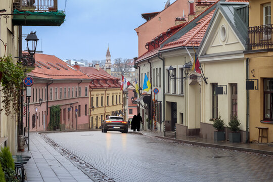 
a narrow, cobbled section of the street with colorful, multi-story houses, on which one car drives, while the city skyline with a tower can be seen in the background.