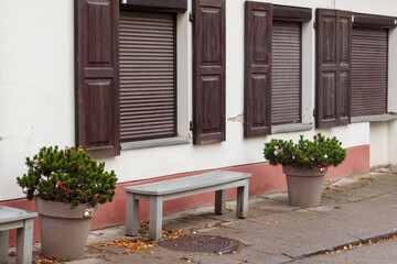 
facade of a light-walled house with several windows closed with blinds, two benches and potted plants are placed next to it.