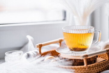 golden black tea in a transparent ribbed cup with a saucer on a wicker tray on a white windowsill, light composition, candies in gold packaging, candle, spring tea, white decor, bouquet
