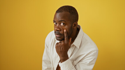 Young man with attentive expression pointing at his eye standing against a vibrant yellow background.