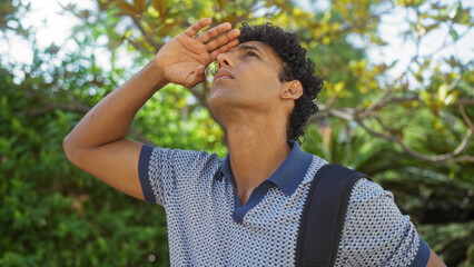 Young hispanic man looking up outdoors in a city park during a sunny day, showcasing urban greenery and casual summer fashion