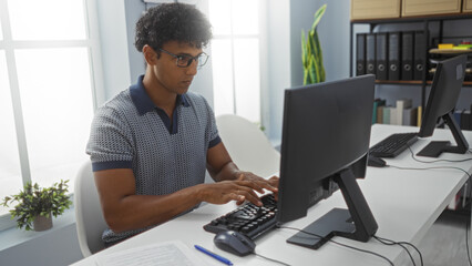 Fototapeta premium Young man working in an office on a desktop computer, with shelves and plants in the background, showcasing a professional workplace environment.