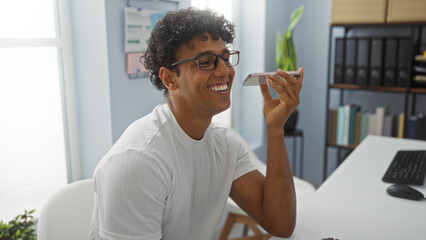 Young man with glasses smiling while using smartphone for voice command in a modern office setting