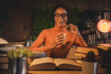 Young woman studying at home enjoying a snack break in cozy evening ambiance surrounded by books