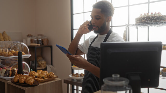 Young man working in a bakery uses mobile phone while holding a tablet, surrounded by pastries and bread in an indoor shop setting, suggesting business and multitasking - Powered by Adobe