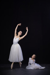 Fototapeta premium Adult ballerina posing with arms elegantly curved above head, child sitting nearby gazing upwards, both in white costumes, against black background. Concept of ballet, elegance and beauty, grace. Ad