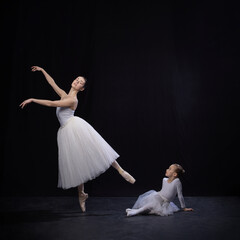 Naklejka premium Adult ballerina posing mid-dance with leg raised, child sitting on floor gazing up, both in white ballet costumes, against black studio background. Concept of ballet, elegance and beauty, grace. Ad
