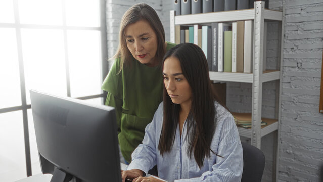 Women working together in an office, with one woman in a green shirt supervising another woman at a computer.