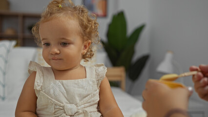 Fototapeta premium Toddler girl with blonde hair sitting on a bed in a bedroom at home, focused on a person feeding her indoors, evoking adorable childhood moments in an interior domestic setting.