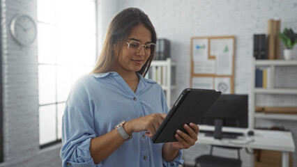 Young woman using tablet in modern office setting, wearing glasses and blue shirt, standing near window with light, working in professional environment with computer in background