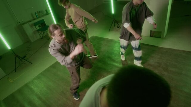 High angle shot of three male Caucasian zoomers and young man improvising and dancing hip hop, moving in circle on dance floor illuminated by green neon lights