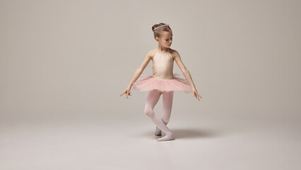 Charming little ballerina kid, wearing pink tutu and tiara, gracefully extending arm while standing on pointe against white studio background. Concept of ballet, elegance and beauty, grace. Ad