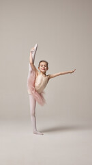 Girl in pink tutu and tights holding extended leg in high split position, smiling confidently showing flexibility against white studio background. Concept of ballet, elegance and beauty, grace. Ad