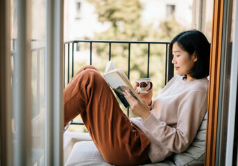 Asian woman relaxing on balcony reading a book with coffee cup in hand
