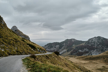 Narrow car road to the Sedlo - Saddle pass, North of Montenegro Durmitor National Park. Calm cloudy autumn landscape scene. Rocky mountain peaks in the clouds. A valley with dried yellow grass.