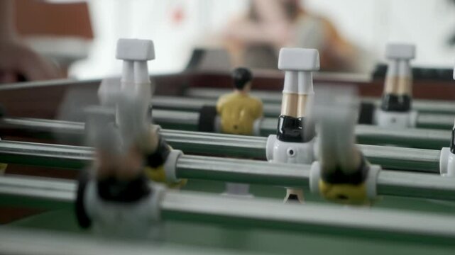 After goal in kicker teenager takes ball out of special hole and puts it into game, static shot. Friends play table football and actively rotate handles with figures of football players.