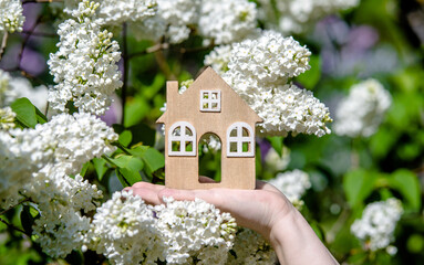 The girl holds the house symbol against the background of blossoming white lilac
