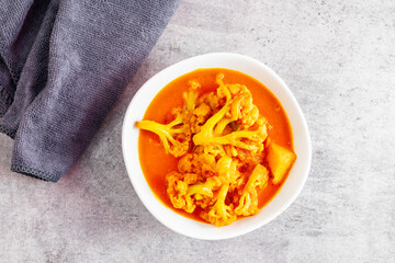 a bowl of vibrant orange-yellow curry with cauliflower florets and potatoes, placed on a Gray textured surface beside a towel.