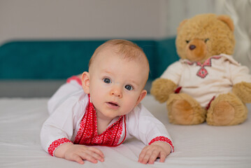 beautiful child boy lying on bed in Ukrainian embroidered shirt with teddy bear toy
