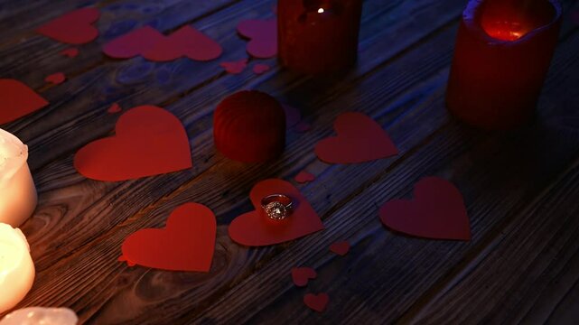 Close up of wooden table in darkness with red and white lighted candles and scattered red paper hearts, diamond ring laying on the heart shaped card with jewelry box.