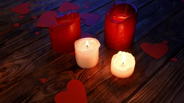 Close up of wooden table in darkness with red and white lighted candles standing on the surface, small red paper hearts scattered around on the desk.