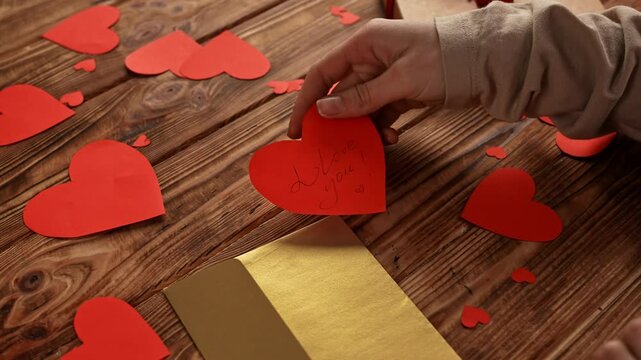 Close up of wooden table scattered red paper hearts on the surface, woman putting heart shaped valentines card with text I love you in golden envelope.