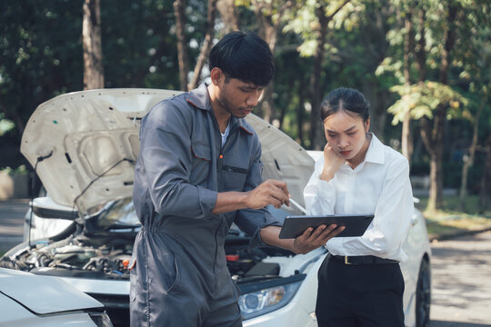 Car Trouble Consultation: A concerned female customer discusses car repair options with a professional male mechanic using a digital tablet, standing by a vehicle with its hood open.