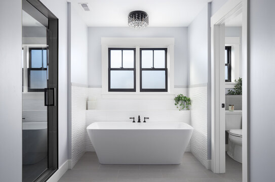 A bathroom detail with a black light fixture above a freestanding soaking tub, shower with a glass door, grey tile flooring, and a wavy white tile backsplash behind the bathtub.