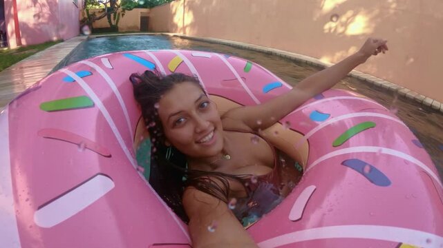 Beautiful girl in a swimsuit floats in a pink inflatable circle in the shape of a donut. Cheerful female smiling through donut inflatable ring. Woman relaxing on donut lilo in the pool water in hot