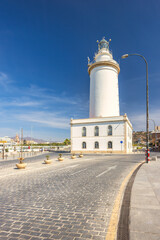 Malaga, seaside city in Andalusia, Spain, Europe. Coastal lighthouse on a sunny day.  A white tower stands tall against a clear blue sky, near a paved road.