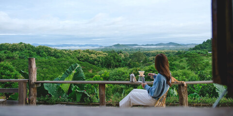 Rear view of a woman making drip coffee while sitting on wooden balcony with a cat and a beautiful mountain view