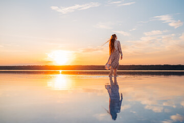 A young woman with long, windblown hair smiles peacefully by a calm lake during a golden sunset,...