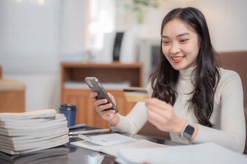 Young asian businesswoman holding credit card and using smartphone making online payment working at home office with pile of paperwork and calculator