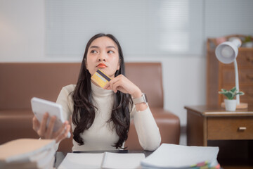 Young asian woman calculating domestic expenses and holding a credit card looking pensive and...