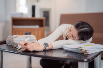 Young asian office worker falling asleep on a desk full of paperwork, holding a pen in her hand and wearing a smartwatch, showing the concept of work stress and burnout