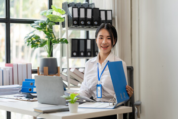 Confident Businesswoman at Desk: A young professional woman sits at her desk, smiling confidently with a blue folder in hand, showcasing dedication and professionalism in a modern office setting.  