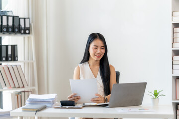 Confident and Focused:  A young businesswoman, radiates professionalism as she reviews documents and works on a laptop at her sleek, organized desk, radiating confidence and focus. 