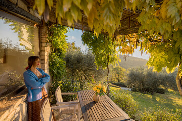 A serene woman standing by a rustic wooden table on a patio surrounded by lush greenery, golden...