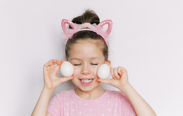A cute and happy little girl in a pink T-shirt with pink ears holding Easter eggs. White background