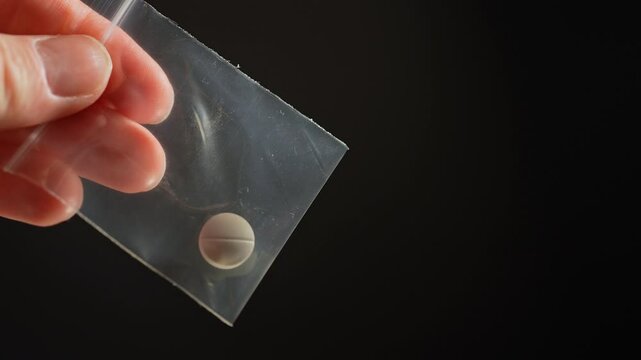 Hand of the drug dealer show off and lure with prescription opioid pill in small plastic bag for sale in clubs, parties and street to addicts. Illegal drugs trafficking, cause dependence and overdose