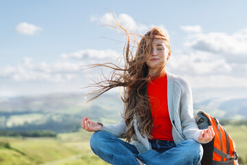 Young woman meditating on a rocky outcrop in a lush green landscape with a backpack nearby