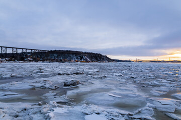 Panoramic blue hour winter view of frozen St. Lawrence river, with cliff and houses along the Chemin de la Plage Jacques-Cartier and bridges in soft focus background, Quebec City, Quebec, Canada