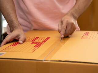 Close-up of man hands cutting through packing tape on cardboard box with red shipping labels,...