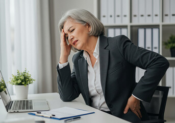 SENIOR BUSINESS WOMAN IN A SUIT SITTING AT A DESK IN THE OFFICE HOLDING HER HAND ON HER BACK AND HEAD AND SUFFERING FROM SEVERE PAIN. NEEDS REST AND MEDICAL ASSISTANCE