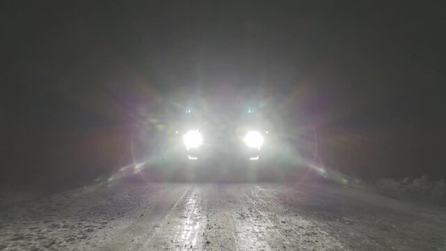 Pickup truck is driving down a snowy road at night. The headlights are on, illuminating the road ahead.