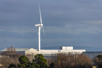 Wind Turbine above Industrial Factory Plant Complex Roofs Against Cloudy Sky and Trees in Foreground